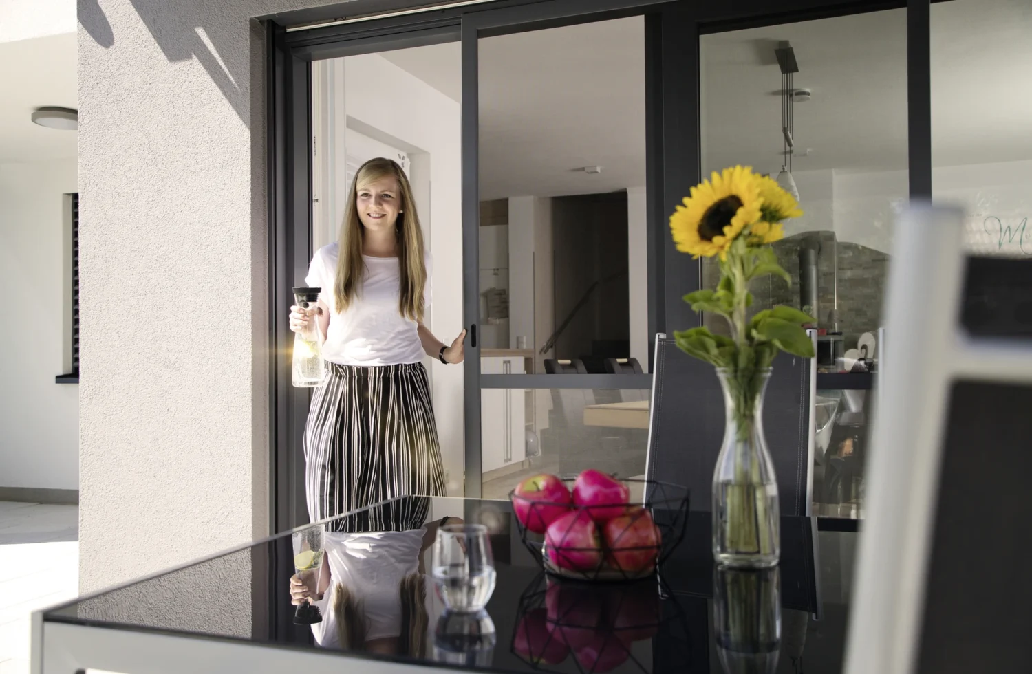 Frau serviert Getränk auf moderner Terrasse mit Sonnenblume und Obstkorb